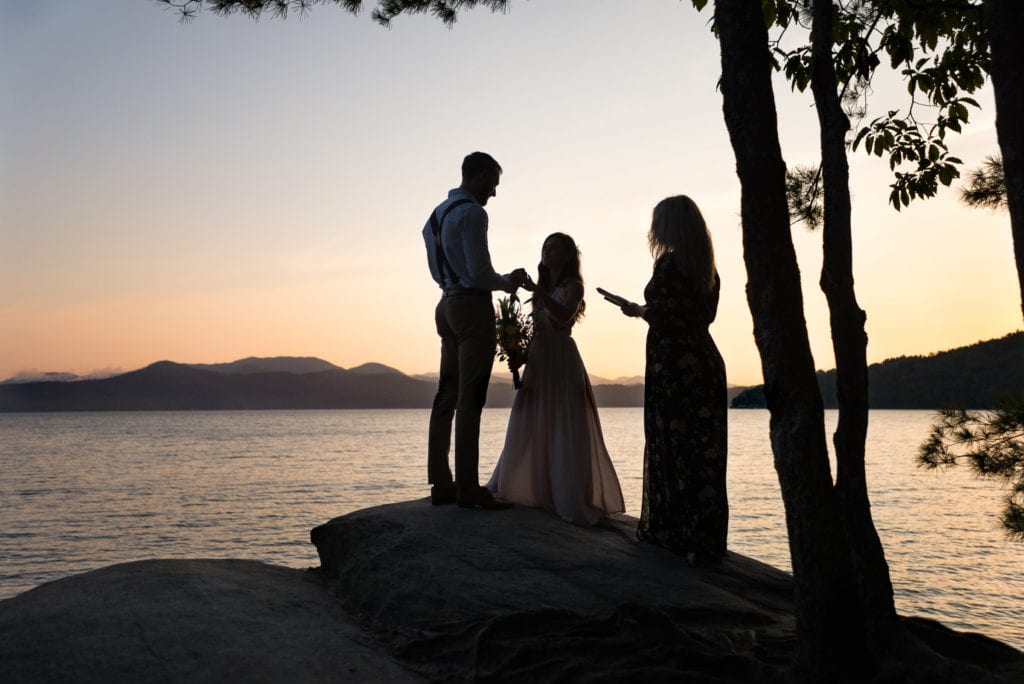 A bride and groom exchange rings in an adventure elopement ceremony on the top of a rock overlook overlooking a South Carolina lake at sunrise.  They chose to elope to break free of a traditional wedding
