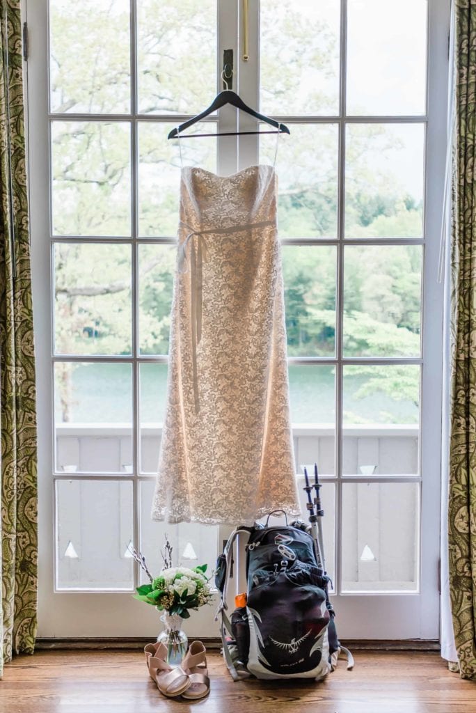With the waters and trees of Lake Toxaway visible through the glass french doors of a bridal suite at Greystone Inn, a the sun shines through a cream-colored lace wedding dress hanging in the doorway. Set below the dress are a backpack with trekking poles for hiking attached to the side, and the bride’s wildflower bouquet and gold colored sandals as the couple readies for their hiking wedding.