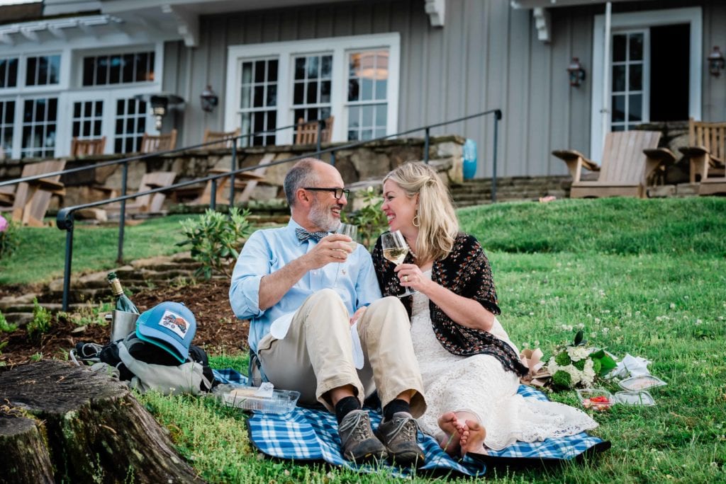 Following their adventure wedding during their stay at Greystone Inn, a barefoot bride and groom with hiking shoes on smile broadly exchanging a champagne toast and picnic on the resort’s lush, green hillside front lawn overlooking Lake Toxaway. The grey wooden building, stone patio with rocking chairs and expansive windows of the Grey Stone Mansion are visible in the background at the top of the hill. This adventure wedding image was taken by MAKE / Adventure Stories Photography.