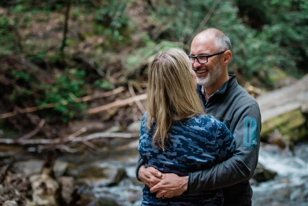 A couple snuggles with big smiles while standing in the creek deep in Pisgah National Forest. The rocky creek is surrounded by lush greenery