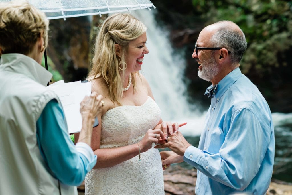 a bride and groom smile happily at each other in a Blue Ridge Mountain waterfall elopement while the groom's mother acts as an officiant. In the background is lush greenery with a gushing waterfall directly behind them in a mountain elopement ceremony.