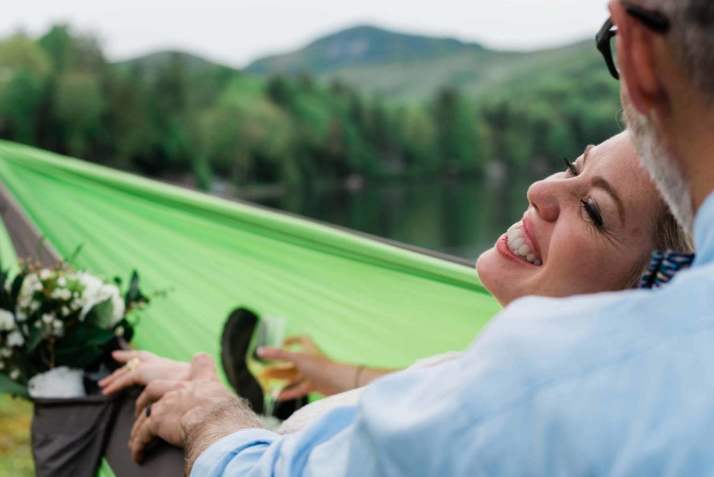 After their waterfall elopement in the Southeast, a bride and groom relax in a hammock together overlooking a beautiful lake, ringed with lush green mountains with trees. The bride leans back into the groom in the hammock in her wedding dress, smiling brightly, while he kisses her head as they relax after their elopement ceremony