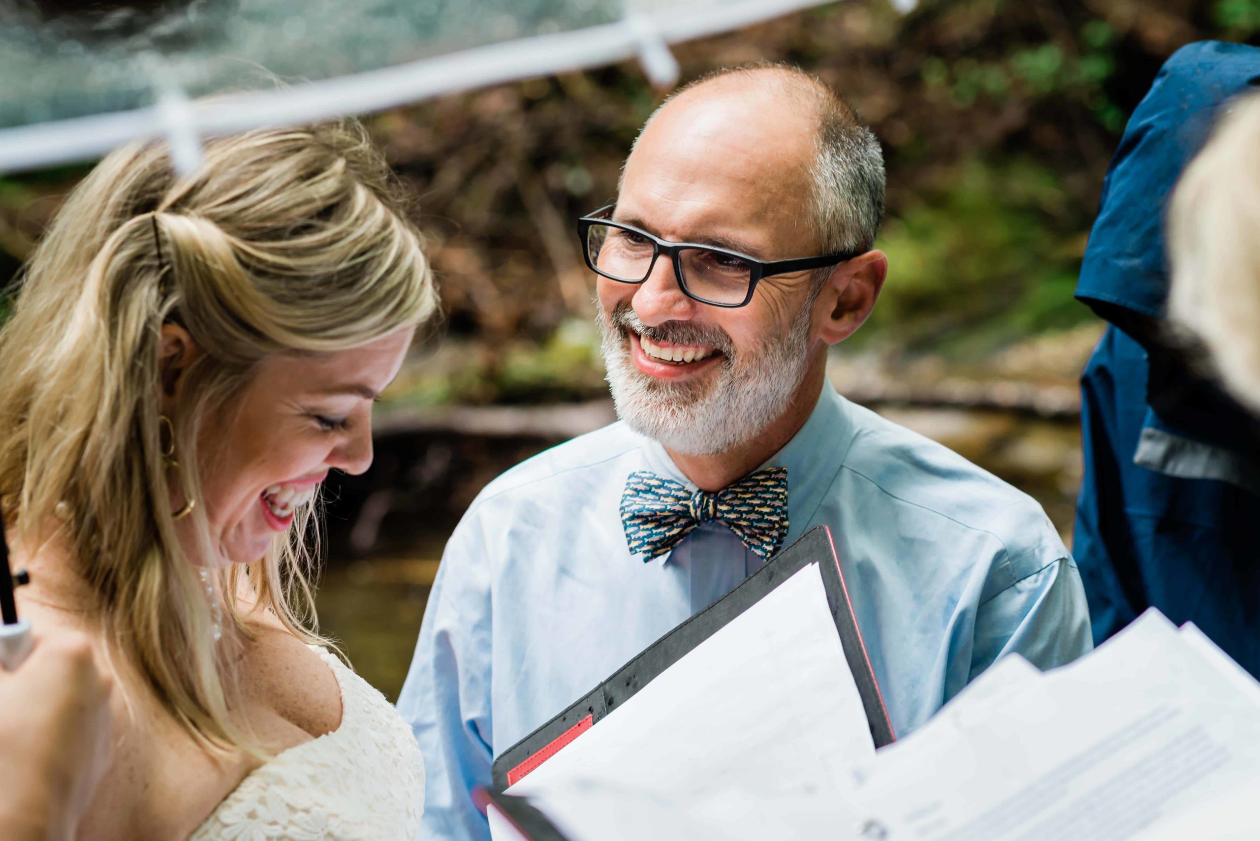 A groom looks at his partner with a huge smile during their hiking elopement ceremony in the rain. They stand together smiling under a clear umbrella.