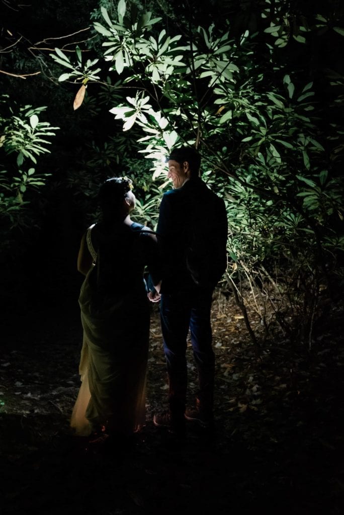 A bride an groom are hiking in the dark through low hanging rhodendron shrubs in an arc overhead