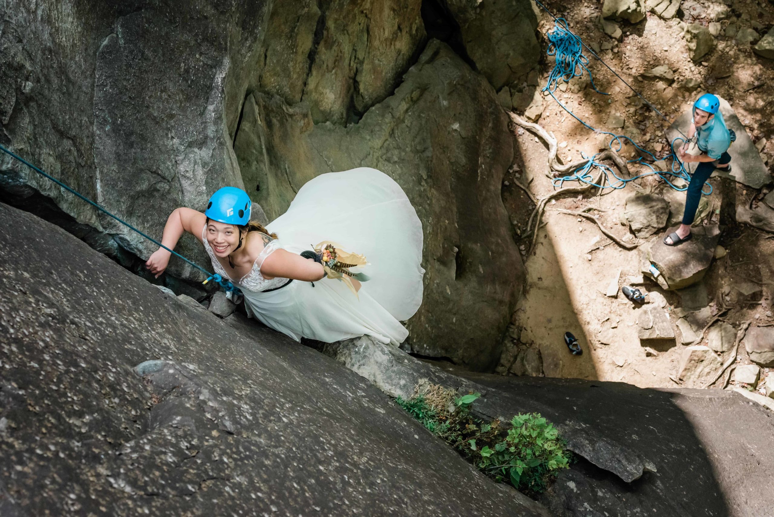A rock climbing bride in a New River Gorge elopement smiles up at the camera. The groom is visible below her as he runs the climbing rope for safety.