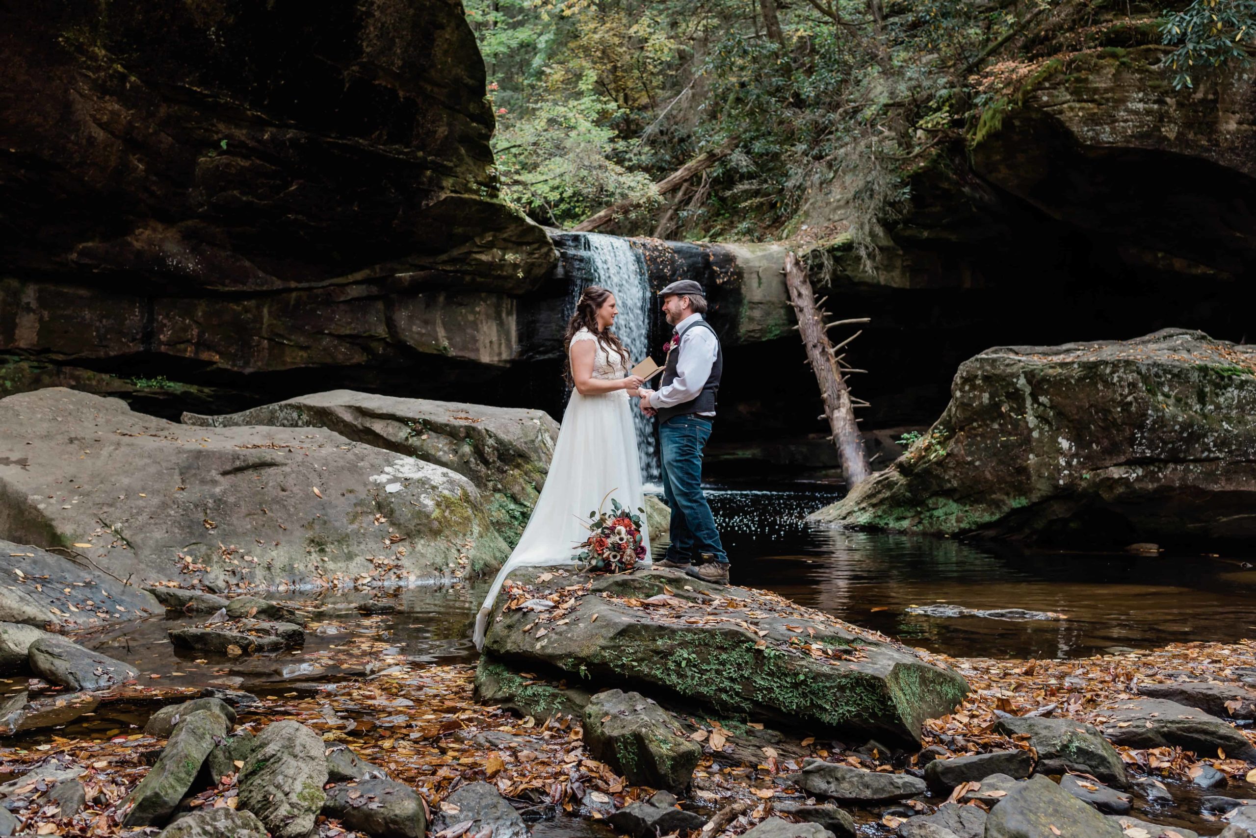in a Kentucky adventure elopement a couple in wedding attire stands on a rock with a water fall behind them The elopement is in a very large natural ampitheatre surrounding them. The bride reads her vows to the groom during this Kentucky hiking elopement surround by rushing water and lush forests and towering rock walls.