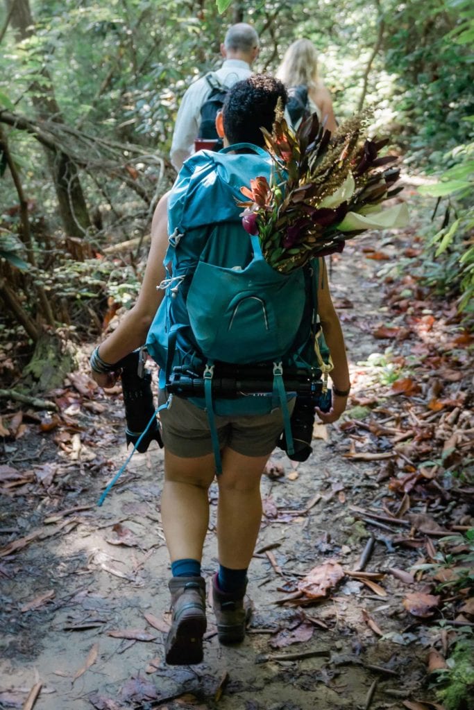 photographer for adventure elopements hikes behind the couple with a large bridal bouquet of flowers sticking out the top of a backpack
