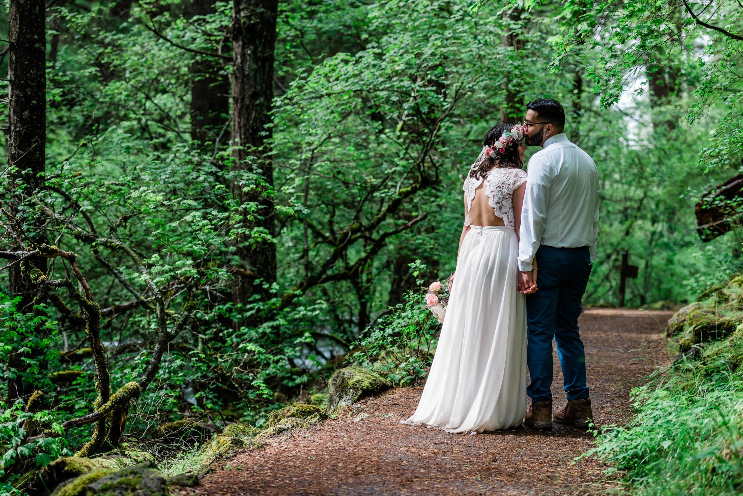 after their waterfall elopement a couple walks on wooded rich green forest path path as the groom kisses the brides head gently