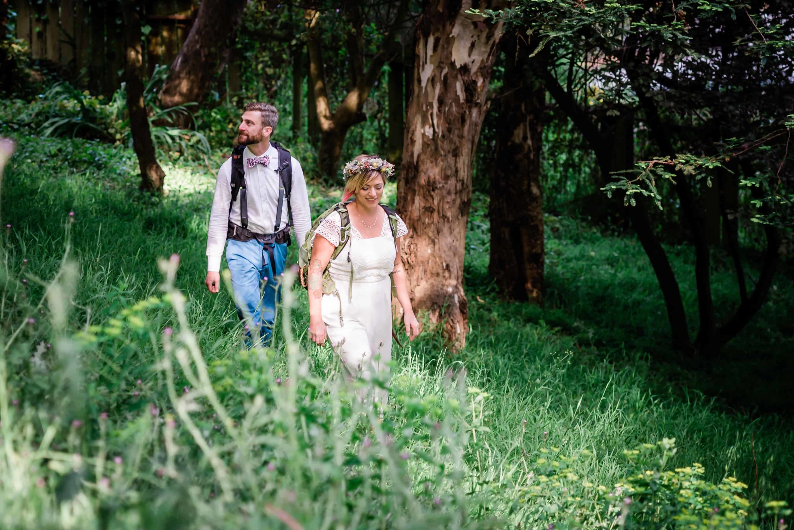 Couple walks through the woods in their elopement attire with backpacks on, admiring the scenery.