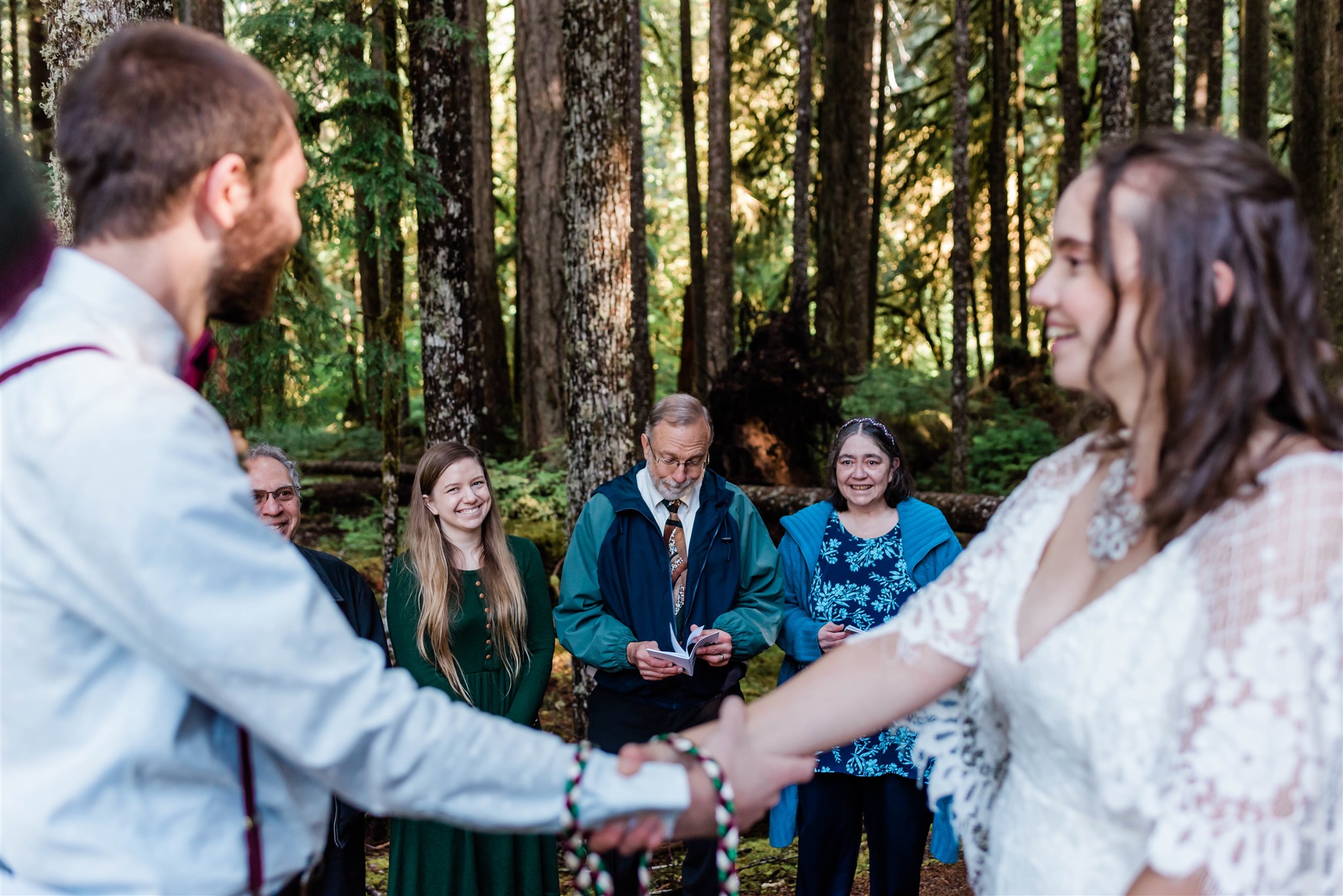 Couple holds hands while family smiles behind them in the forest.
