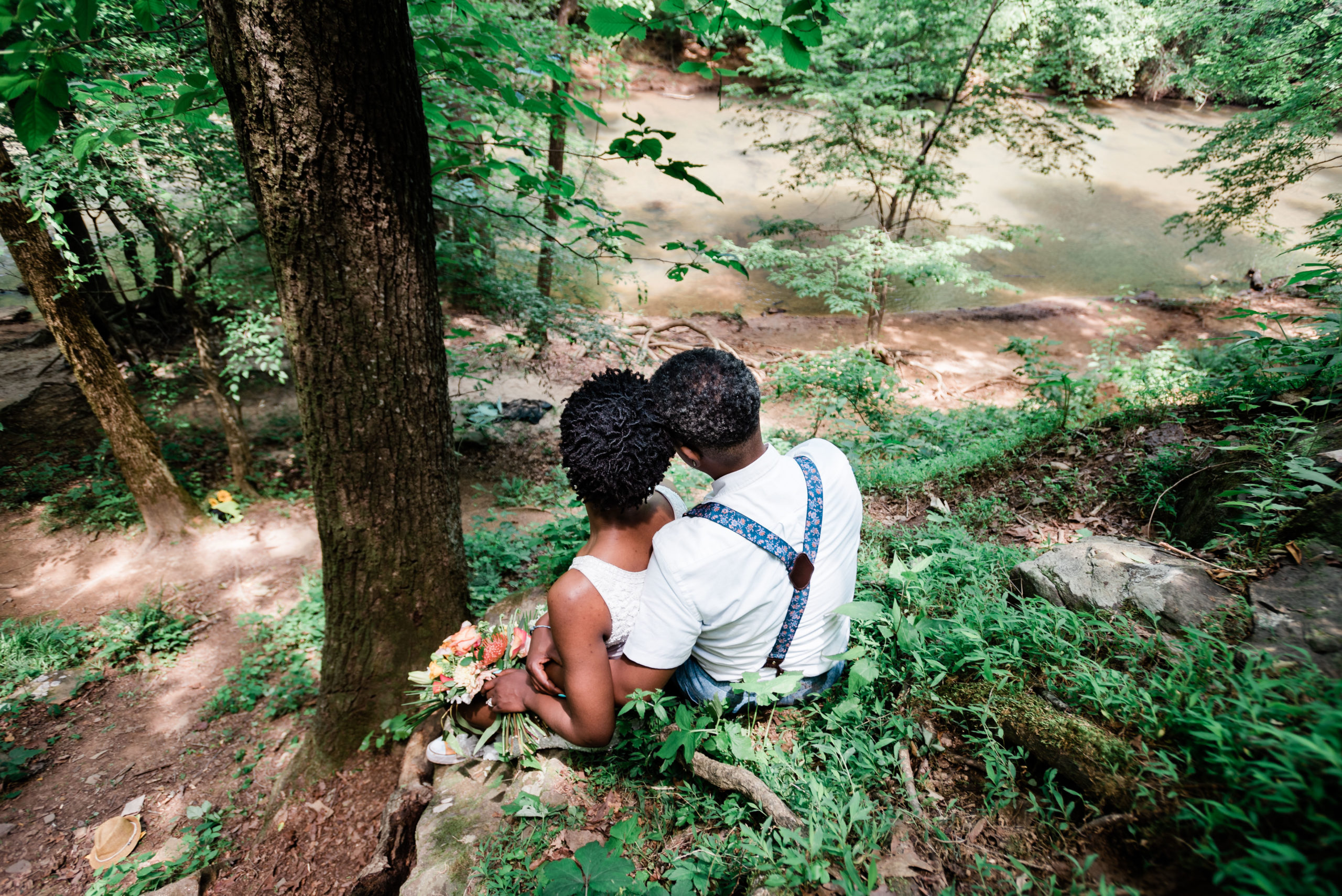 An eloping couple sits on the edge of a dliff in their formal wear, looking away from the camera down to the greenery surrounding a river