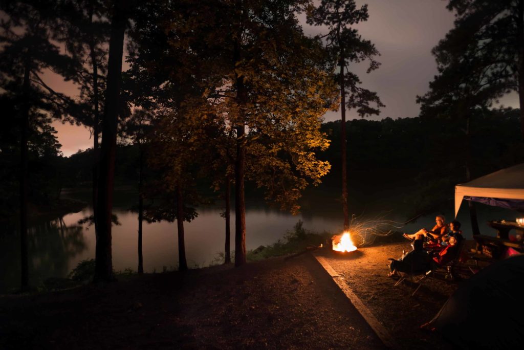 This is a nighttime picture of people sitting next to a lake in front of a bonfire.