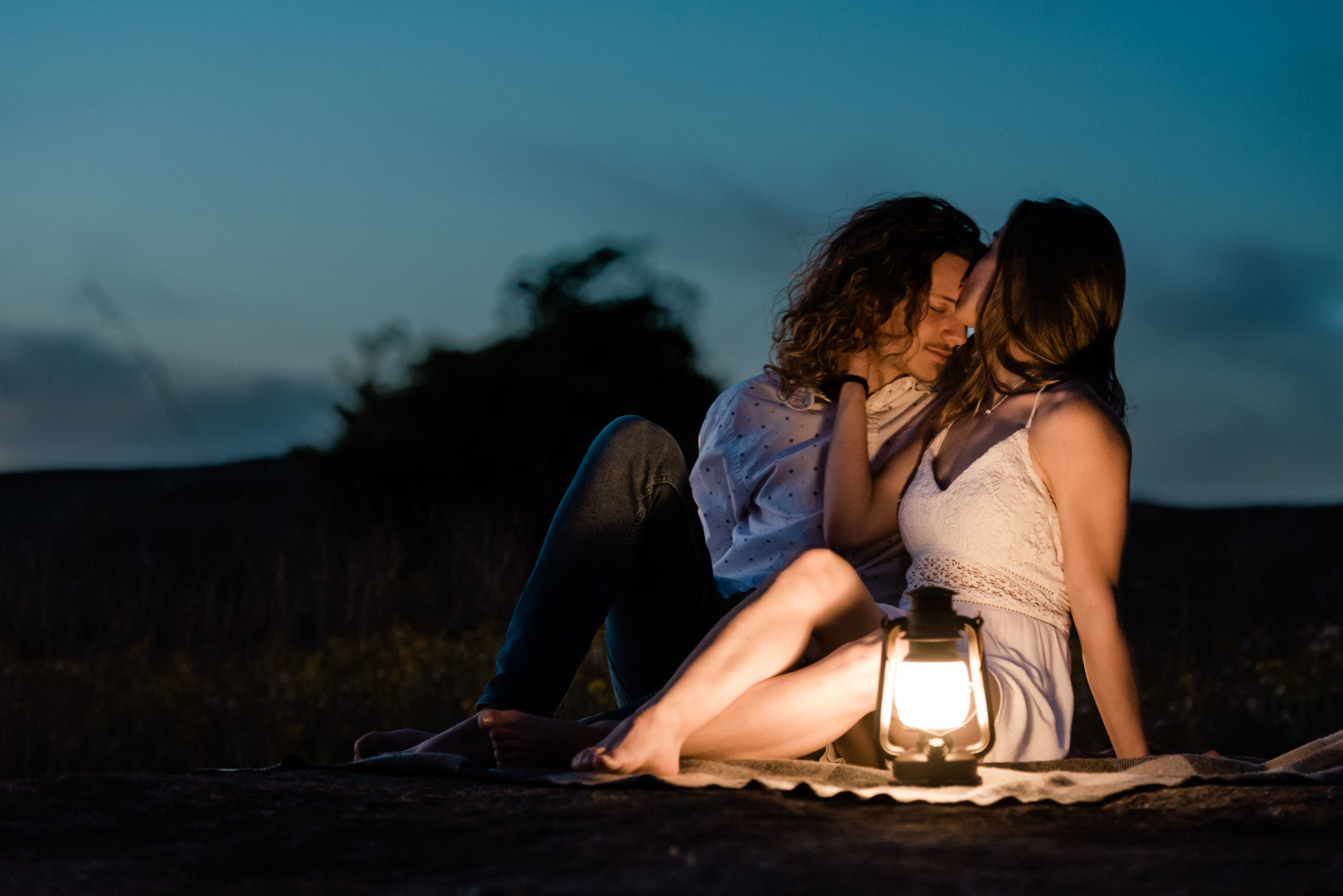 In this picture we see an engaged couple kissing while sitting on a blanket with a lantern. The sun has recently set and it's getting dark outside.