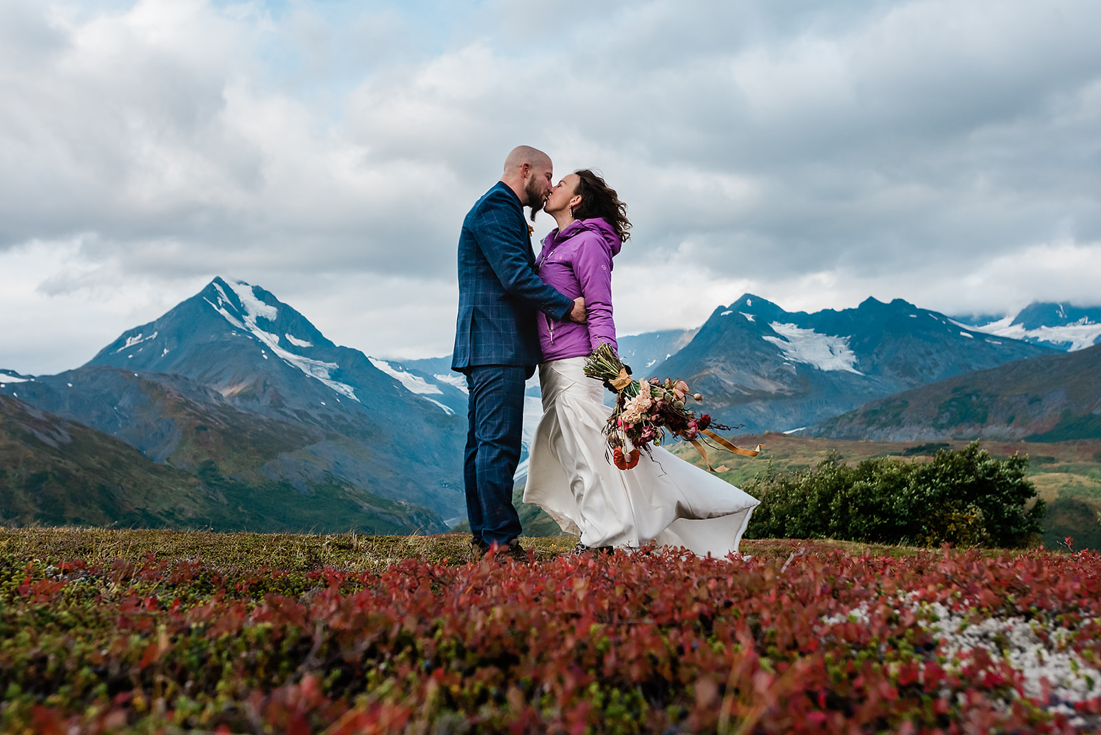 This picture is of a couple standing on a beautiful Alaskan hill with the mountains behind them. They are kissing and is their wedding attire. This couple decided to elope and have an adventure for their wedding experience.