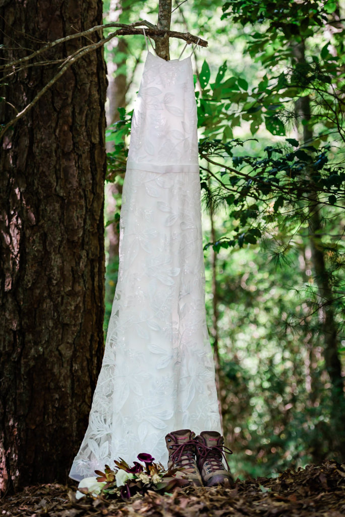 This picture is a white lace wedding dress hung up on a tree in the Georgia Mountains. In front of the dress is a pair of hiking boots. These are a little details for before an adventure elopement.