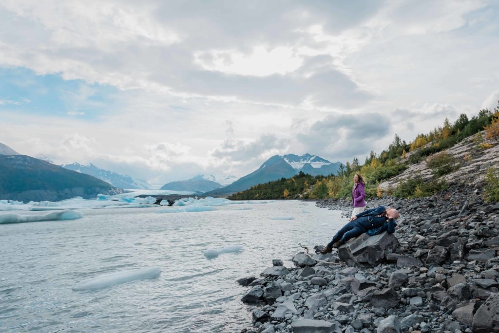after deciding where and how to elope in Alaska, a couple in formal wedding clothes laughs on a rocky shore of a lake with little icebergss and alaska snow capped mountains in the distance