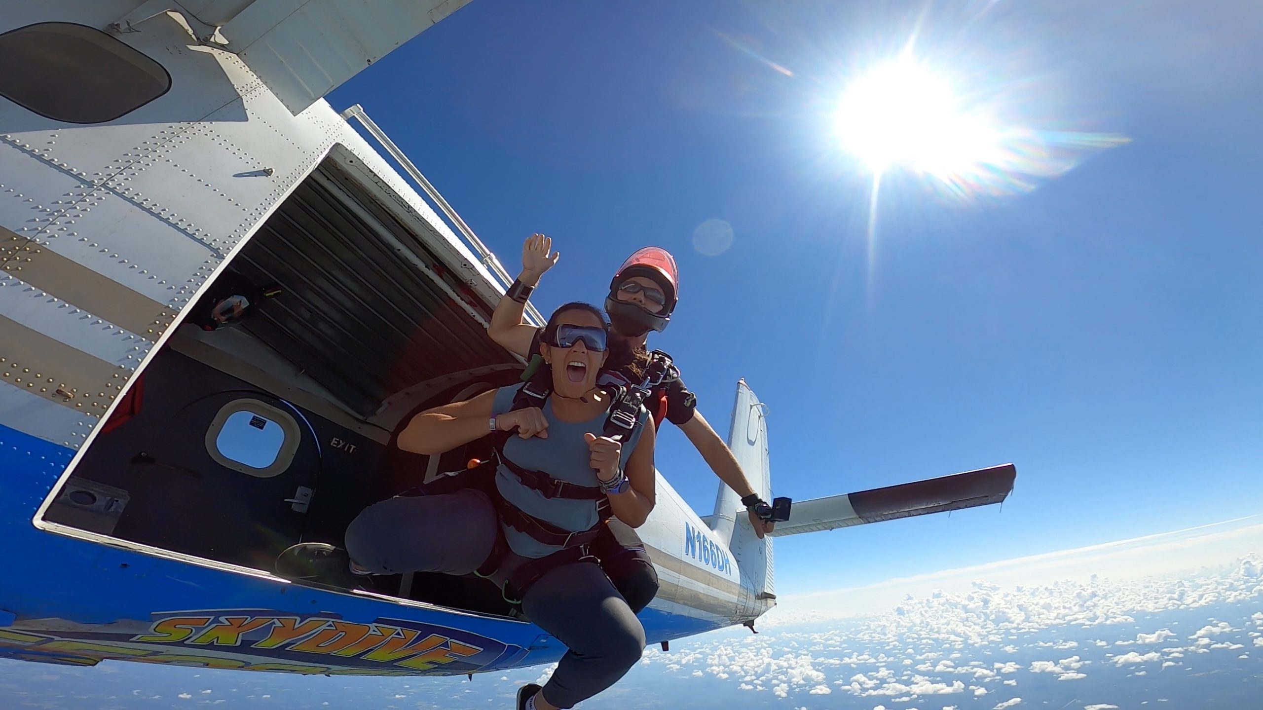 black woman just jumped out of a plane attached to an instructor in a tandem skydive at Skydive Georgia. She is screaming happily in the blue skies with sun shining bright