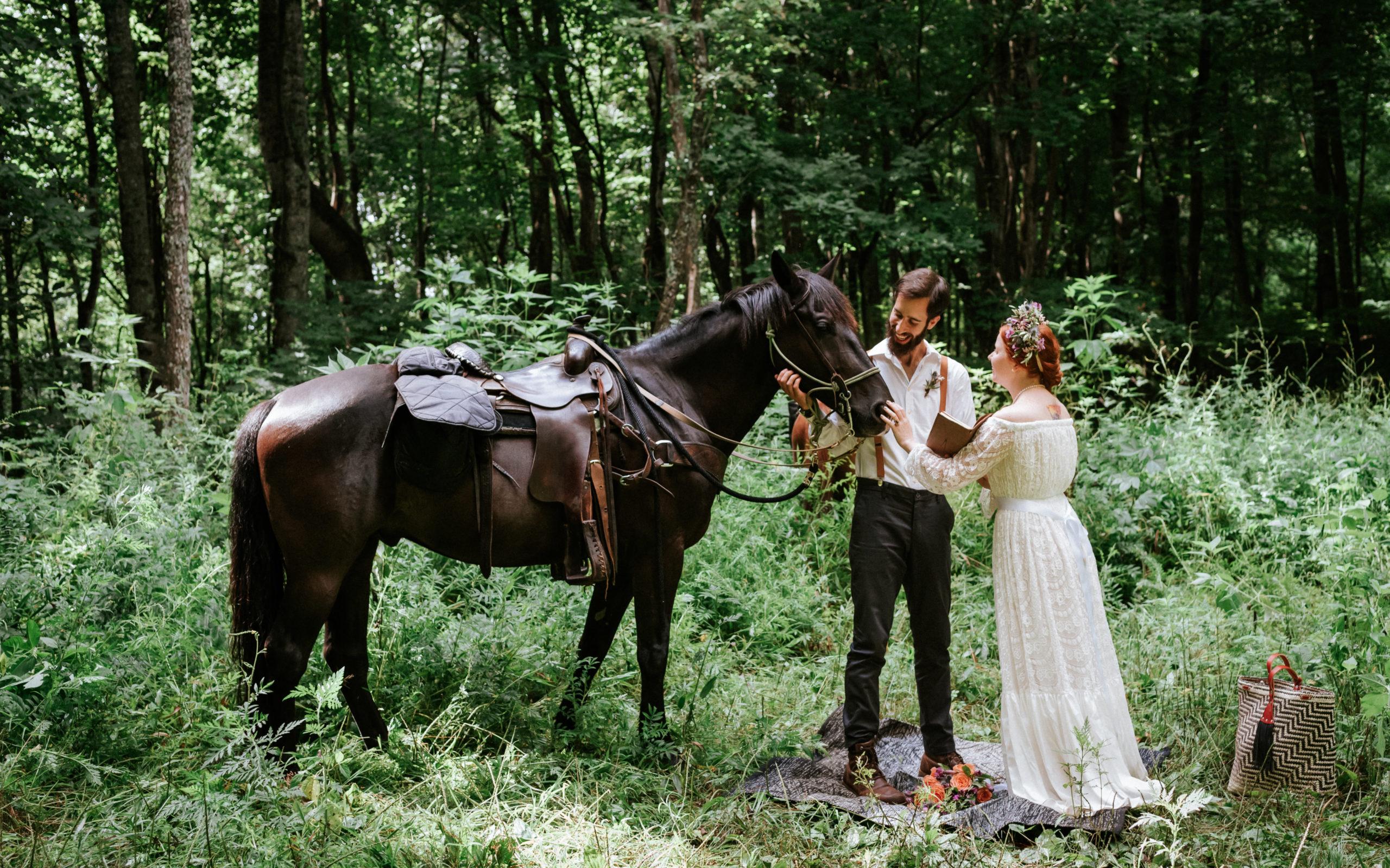 This picture is of a bride and groom petting a horse together. They are in the forest, surrounded by green.