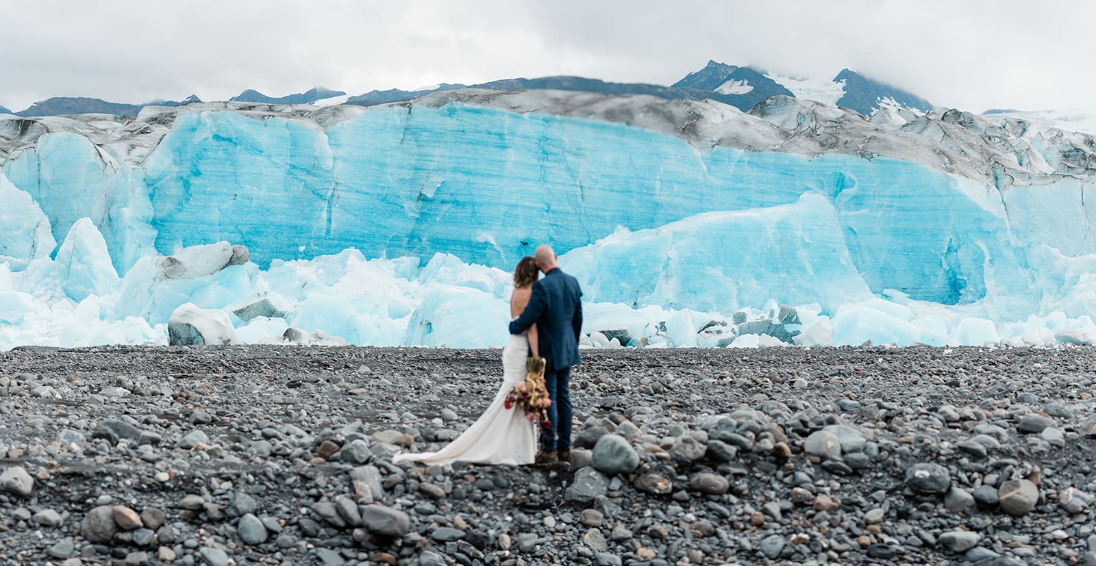 This is a picture of a mountain with a big glacier in front. There is a couple holding each other in front of it.