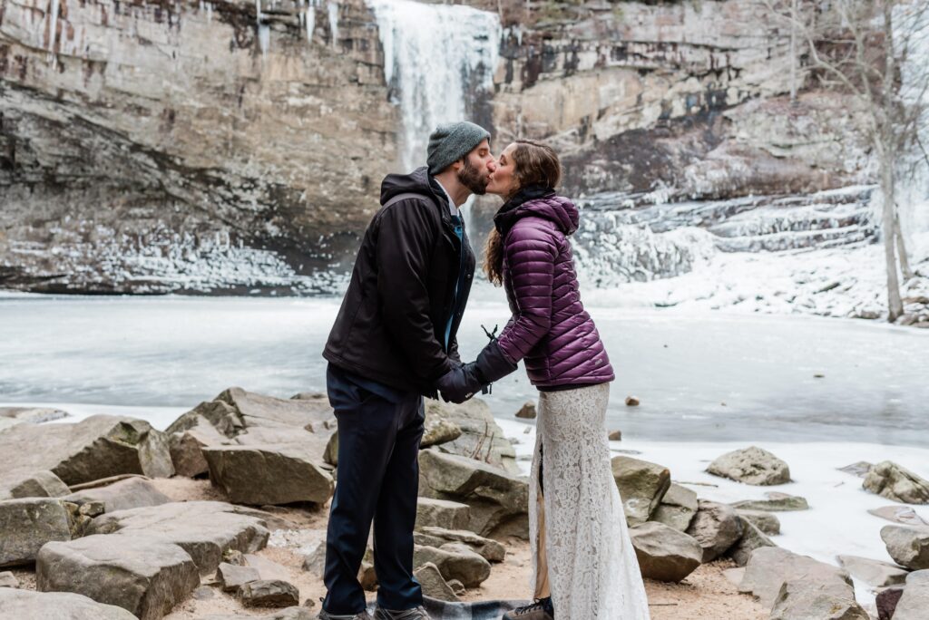 This is a Chattanooga elopement in winter. This picture is the bride and groom bundled up in the snowy forest. They are kissing with a frozen waterfall behind them.