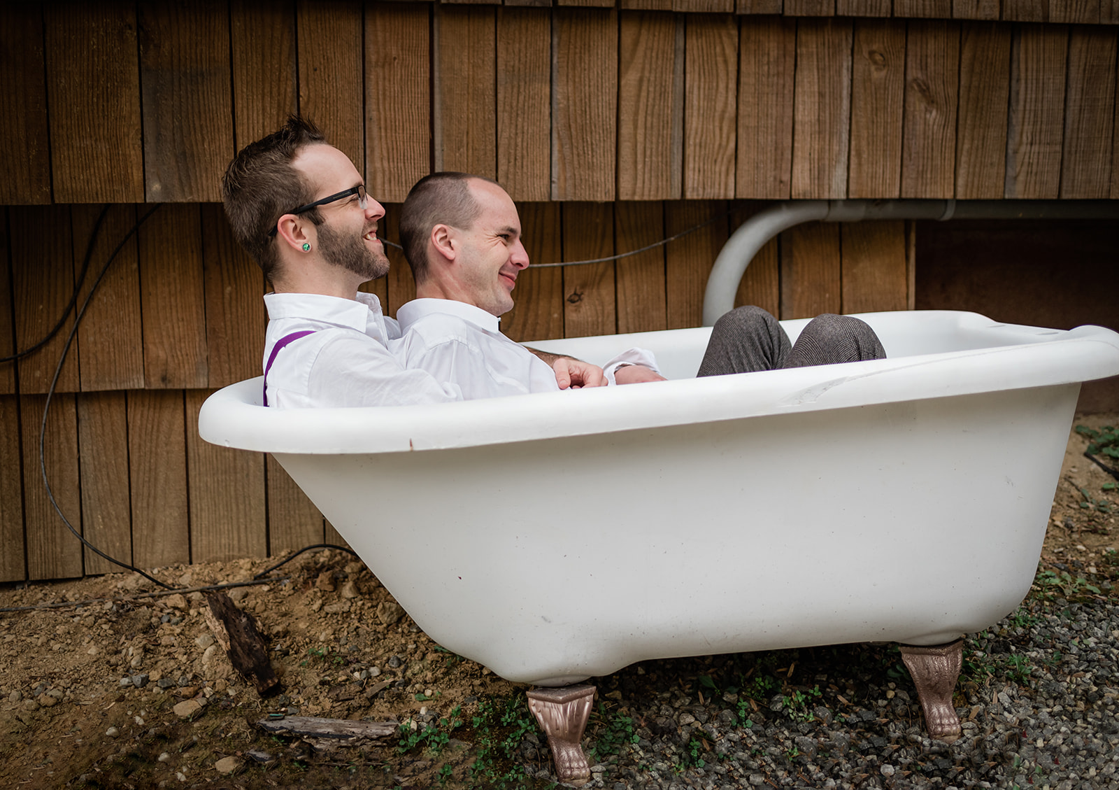 This is a picture of two men laying in a tub together outside. They are fully dressed and laughing together.