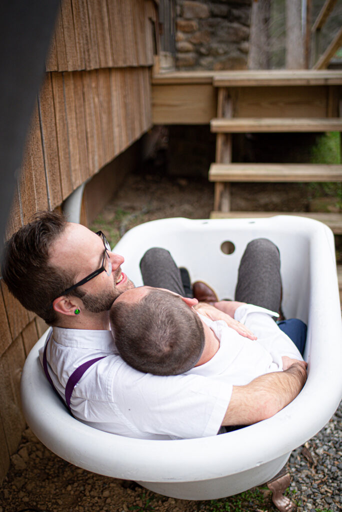 This is a picture of two men laying in a tub together outside. They are fully dressed and laughing together.