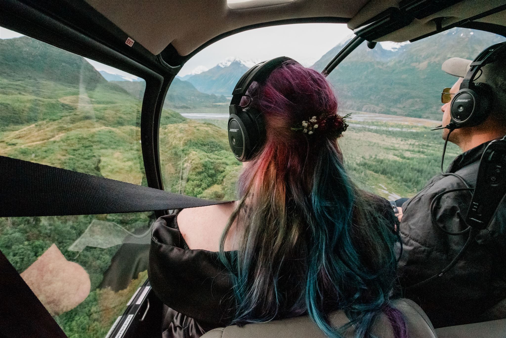 This is a picture of a bride riding in a helicopter. She is sitting in the front looking out at the vast mountain view. You can see the captain next to her flying the helicopter.