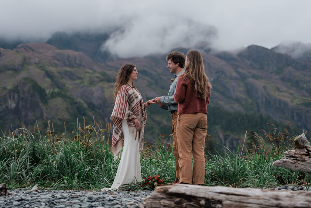 This is a picture of a bride and groom during their Alaskan elopement. They are exchanging rings and there is a foggy mountain behind them.