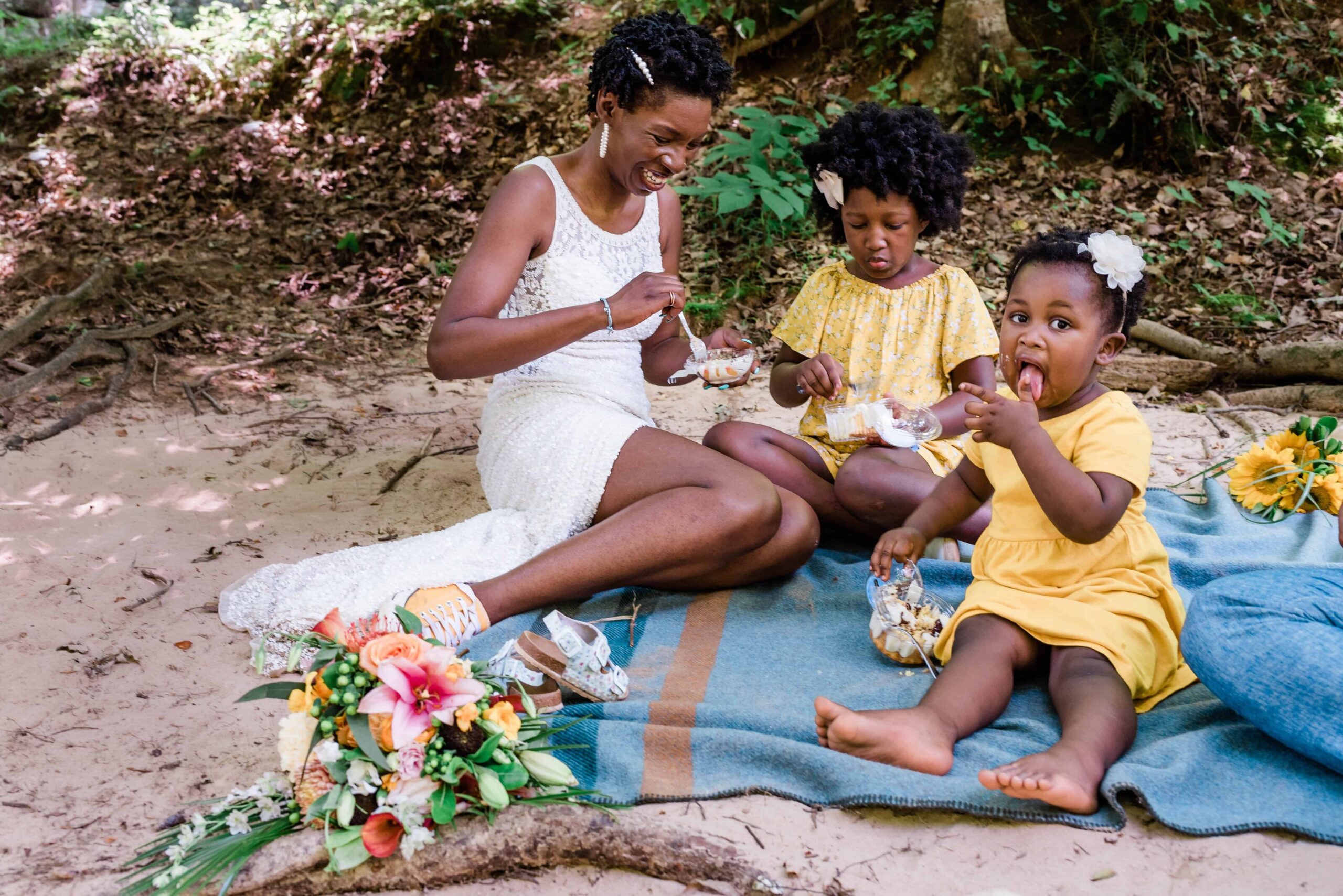 a bride who booked an elopement concierge and photography packages in tennessee and geogria sits on a blanket in her wedding dress eating small wedding cakes with her two young children.